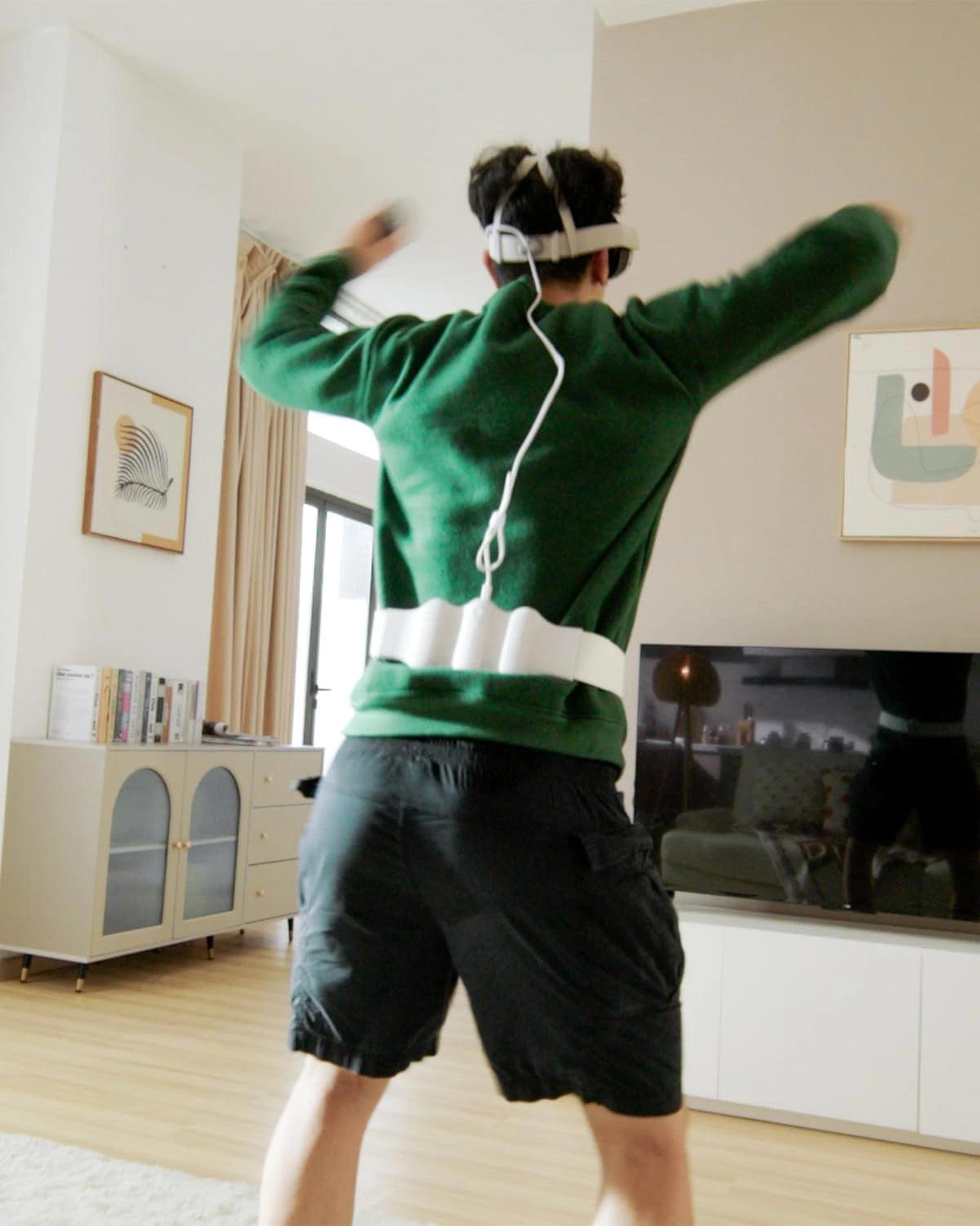 A person wearing a VR headset and green sweater is standing in a living room with arms raised, appearing to engage in a VR gaming experience enhanced by various VR accessories.